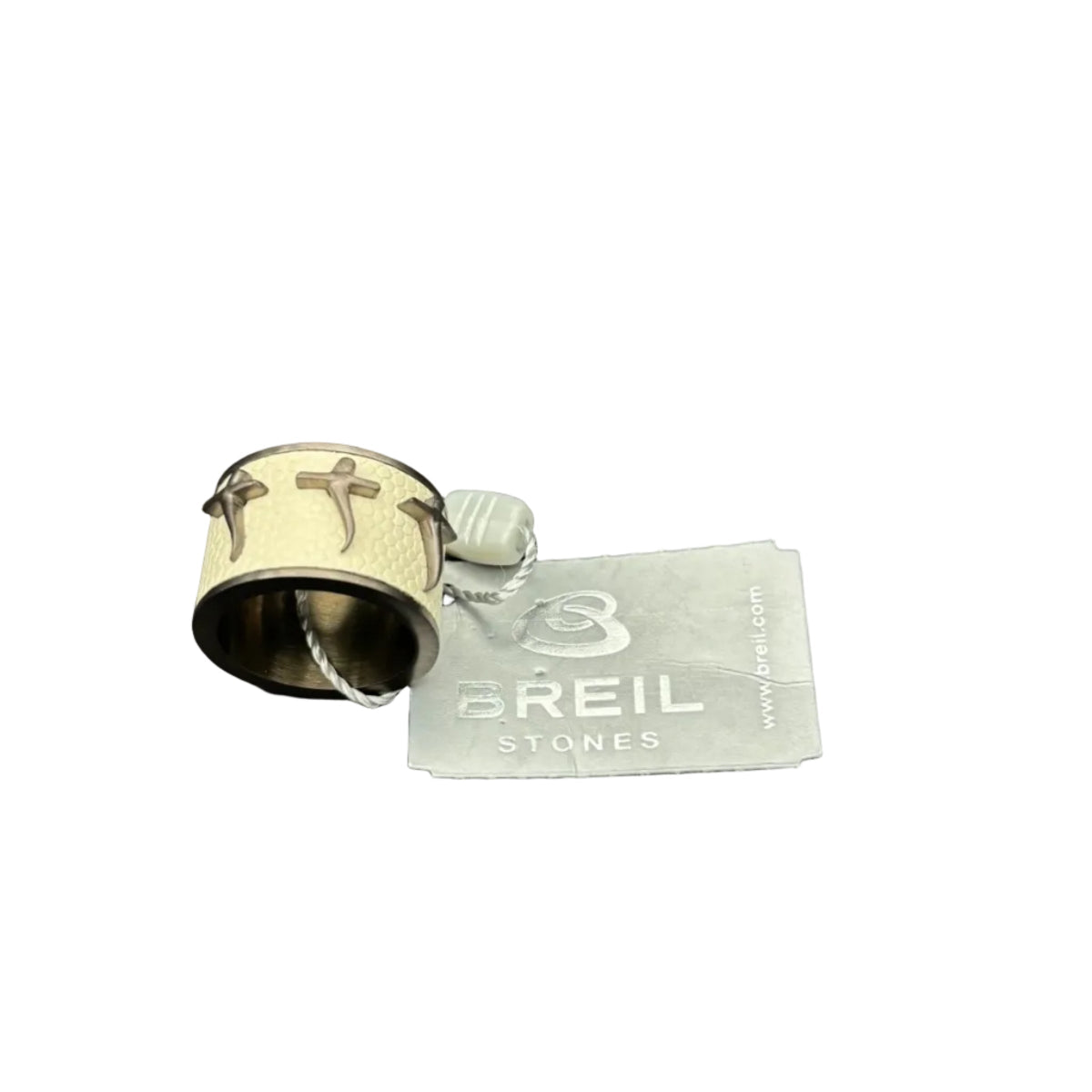 Primo piano di un anello da uomo Breil Stones mostrato da un'angolazione inclinata. Il gioiello presenta una fascia centrale in ecopelle o texture nido d'ape color crema, decorata con tre croci stilizzate. La montatura è in acciaio color bronzo con interno liscio visibile. Sullo sfondo, l'etichetta originale Breil Stones grigia su sfondo bianco.