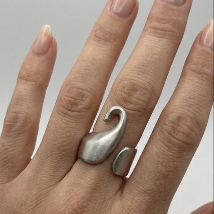 Primo piano di una mano femminile con pelle chiara e unghie con smalto nude, che indossa l'anello Breil in acciaio satinato sull'anulare. L'anello, con il suo distintivo design a forma di cigno stilizzato e fascia aperta, è ben visibile e a fuoco. Lo sfondo è grigio chiaro neutro.