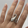 Primo piano di una mano femminile con pelle chiara e unghie con smalto nude, che indossa l'anello Breil in acciaio satinato sull'anulare. L'anello, con il suo distintivo design a forma di cigno stilizzato e fascia aperta, è ben visibile e a fuoco. Lo sfondo è grigio chiaro neutro.