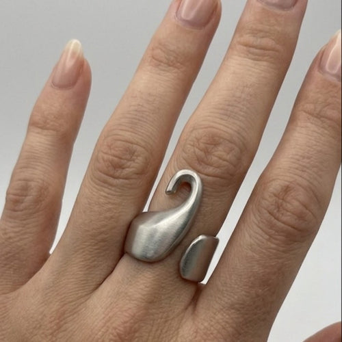Primo piano di una mano femminile con pelle chiara e unghie con smalto nude, che indossa l'anello Breil in acciaio satinato sull'anulare. L'anello, con il suo distintivo design a forma di cigno stilizzato e fascia aperta, è ben visibile e a fuoco. Lo sfondo è grigio chiaro neutro.