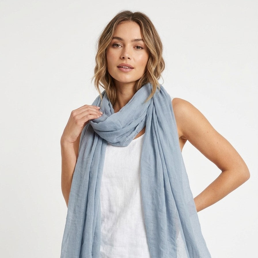 Una modella indossa un leggero foulard estivo di colore azzurro polvere, annodato morbidamente intorno al collo e lasciato cadere sul davanti. Il tessuto appare sottile e leggermente stropicciato, ideale per la stagione calda. La modella indossa un top bianco semplice e posa su uno sfondo bianco neutro e luminoso.