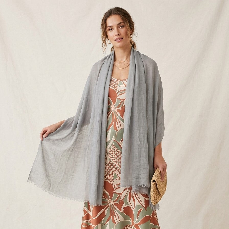 Una modella indossa un ampio foulard estivo di colore grigio chiaro, appoggiato sulle spalle e lasciato cadere lungo i fianchi. Il foulard è realizzato in un tessuto leggero con bordi sfrangiati. L'outfit include un abito lungo con una vivace fantasia floreale nei toni del ruggine, verde e panna. La modella tiene in mano una pochette in paglia intrecciata e posa davanti a uno sfondo in tessuto panna leggermente drappeggiato.