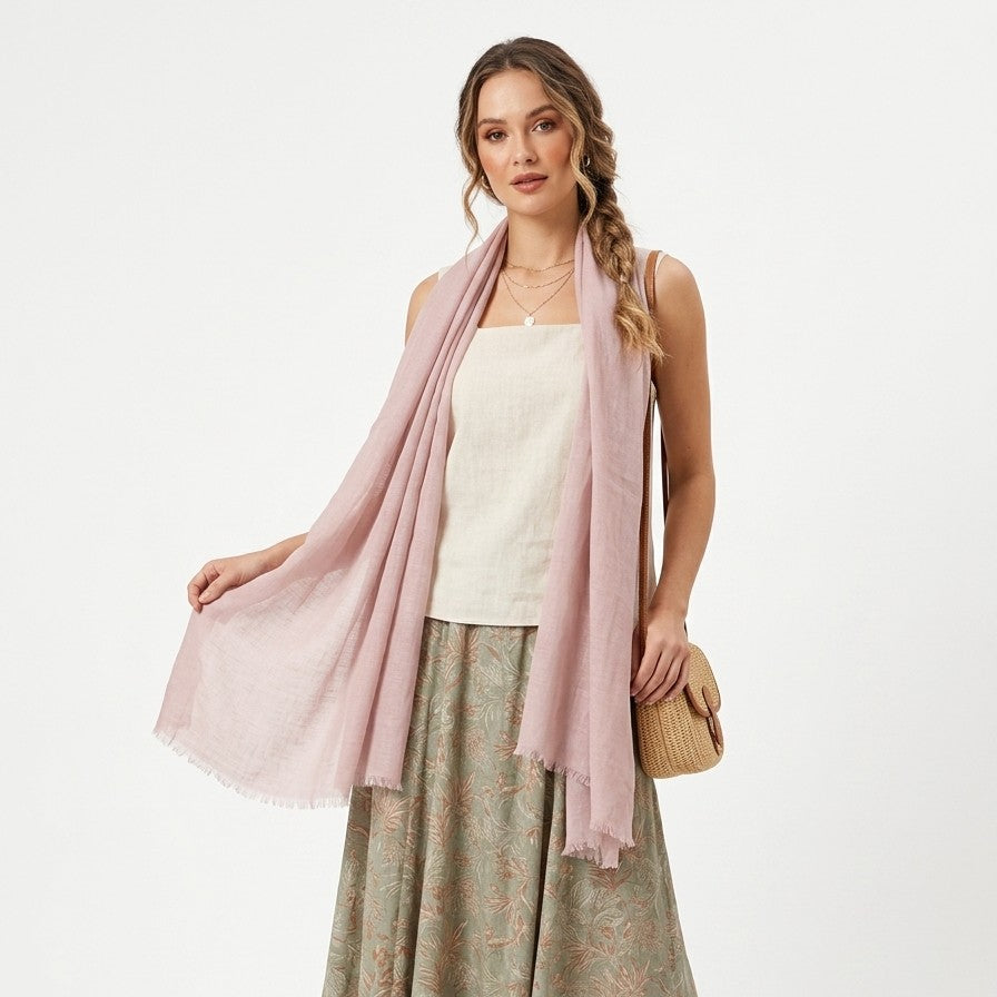 Una modella indossa un ampio foulard di colore rosa antico leggermente trasparente, appoggiato morbidamente sulle spalle. Il foulard presenta bordi finemente sfrangiati. Lo sfondo è bianco uniforme.