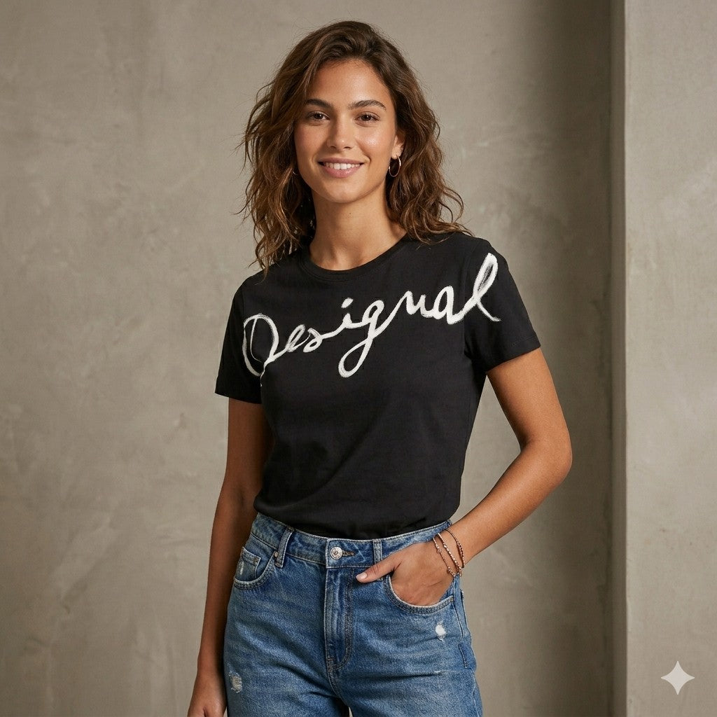 Una modella sorridente indossa una maglietta nera a maniche corte con la scritta 'Desigual' bianca sul davanti. La modella completa l'outfit con un paio di jeans blu a vita alta, piccoli orecchini a cerchio e dei braccialetti sottili. Posa con una mano in tasca davanti a una parete materica grigia dal design moderno.