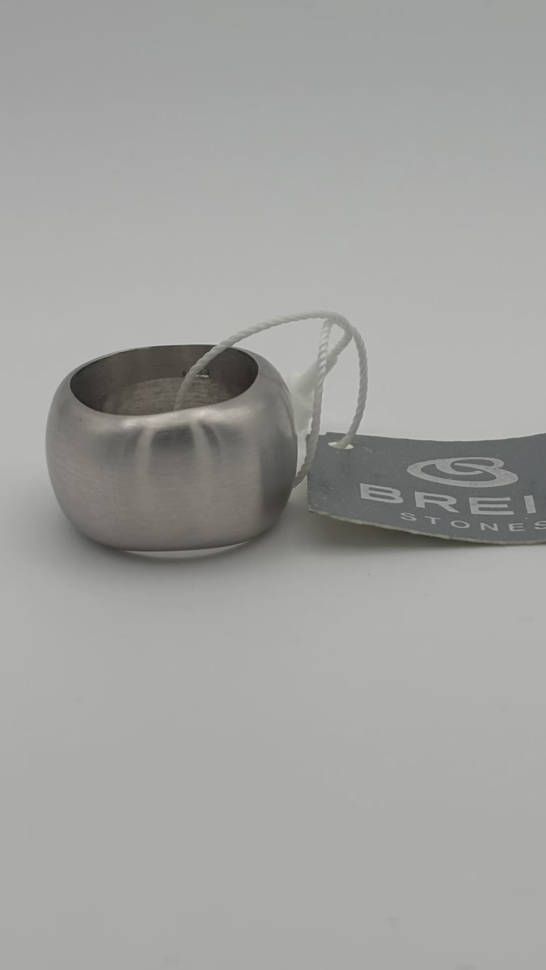 Primo piano di un anello a fascia larga in metallo satinato con etichetta 'Breil Stones'. Una lente d'ingrandimento circolare è sovrapposta alla base dell'anello per evidenziarne la finitura e la superficie liscia.
